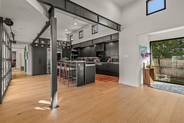 kitchen with light wood finished floors, tasteful backsplash, a breakfast bar, dark cabinetry, and a high ceiling