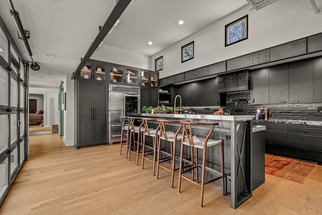 kitchen with an island with sink, light wood-style floors, stainless steel built in fridge, and a towering ceiling