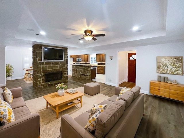 living area with a ceiling fan, baseboards, dark wood finished floors, a tray ceiling, and a stone fireplace