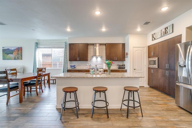 kitchen with light wood finished floors, decorative backsplash, appliances with stainless steel finishes, a kitchen bar, and wall chimney exhaust hood