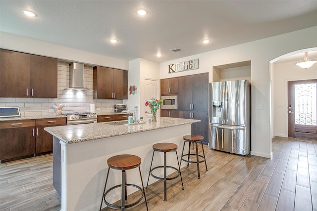 kitchen with a kitchen bar, a sink, tasteful backsplash, appliances with stainless steel finishes, and wall chimney range hood