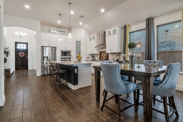 kitchen featuring wall chimney range hood, wood tiled floor, appliances with stainless steel finishes, arched walkways, and a kitchen island with sink