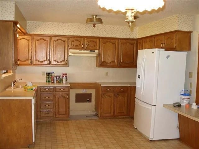 kitchen featuring under cabinet range hood, brown cabinets, wallpapered walls, and white refrigerator with ice dispenser