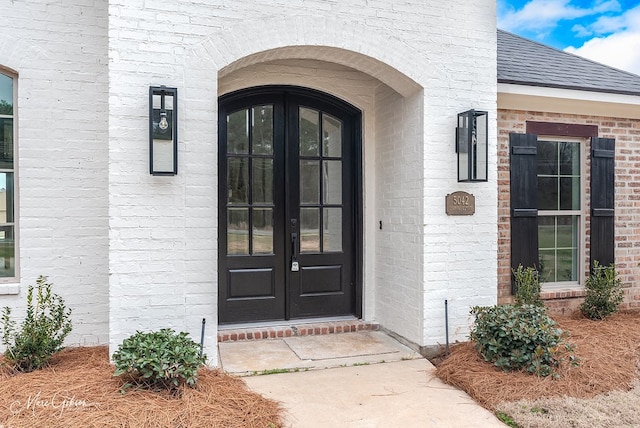 doorway to property featuring brick siding, french doors, and a shingled roof