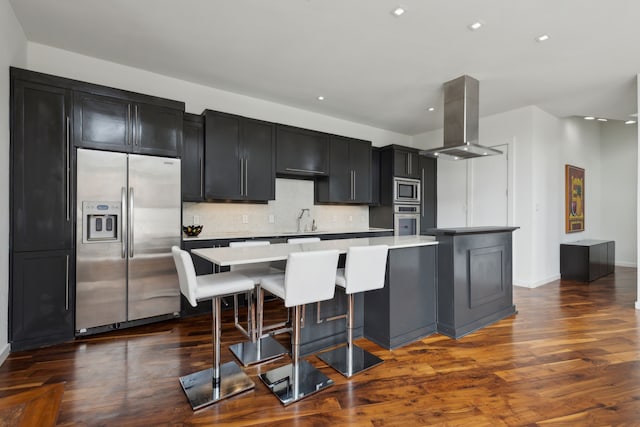 kitchen featuring dark cabinets, stainless steel appliances, a large island, tasteful backsplash, and island range hood