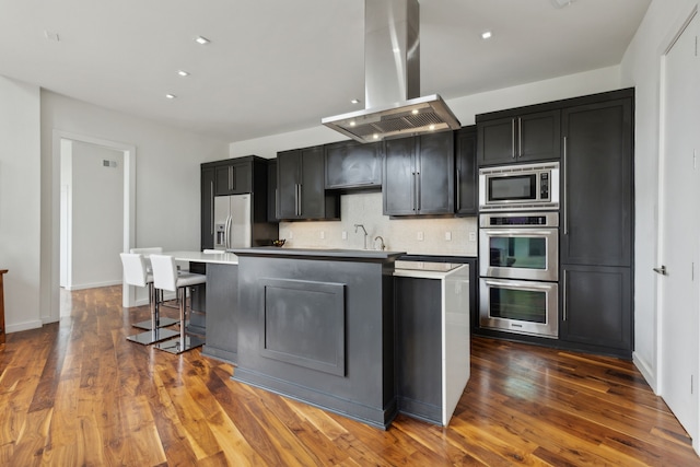 kitchen with a center island with sink, dark wood-style flooring, decorative backsplash, appliances with stainless steel finishes, and island range hood