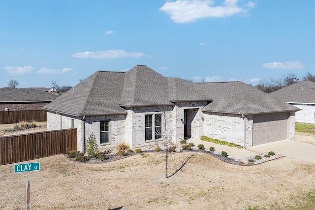 french country style house featuring driveway, fence, brick siding, and roof with shingles