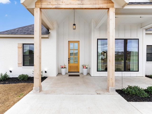 view of exterior entry with brick siding, a patio area, board and batten siding, and a shingled roof