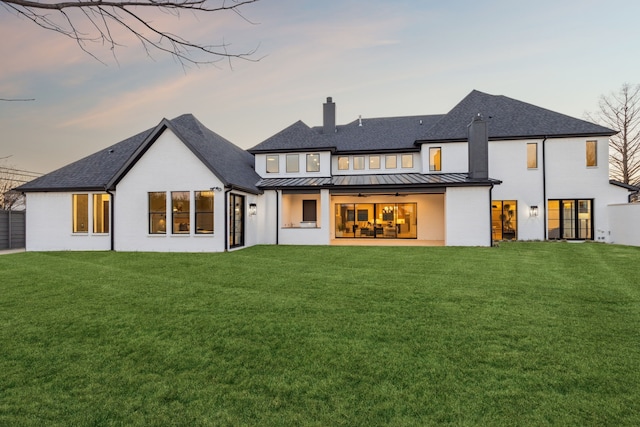 back of house at dusk with a standing seam roof, a chimney, a patio area, a lawn, and metal roof