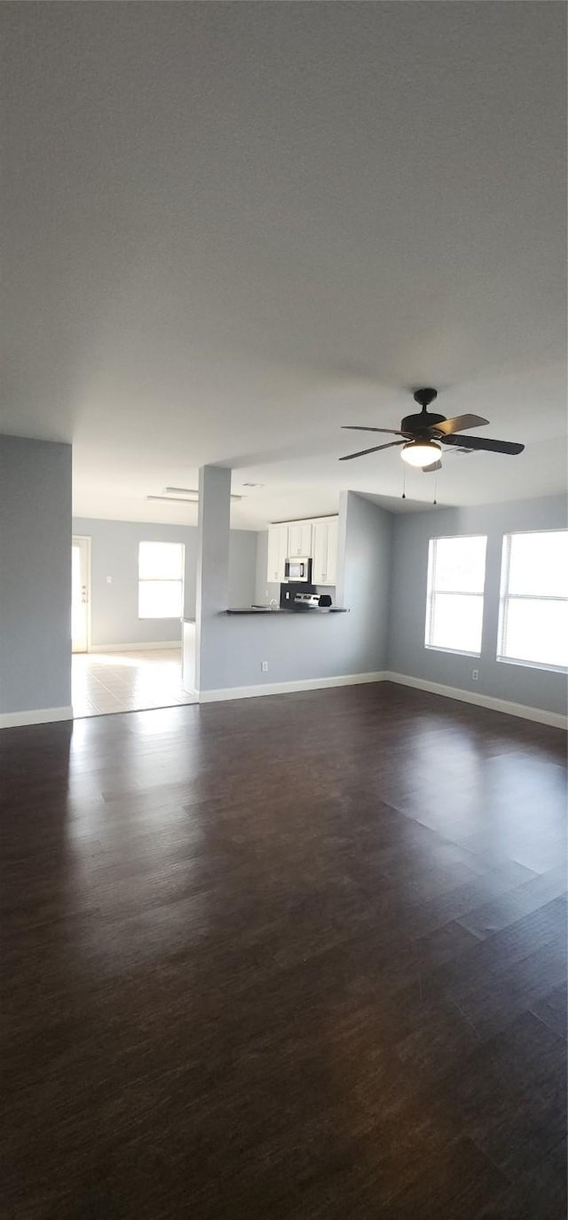 unfurnished living room featuring plenty of natural light, baseboards, dark wood-style flooring, and ceiling fan