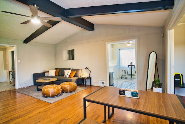 living room with a ceiling fan, wood finished floors, visible vents, lofted ceiling with beams, and a decorative wall