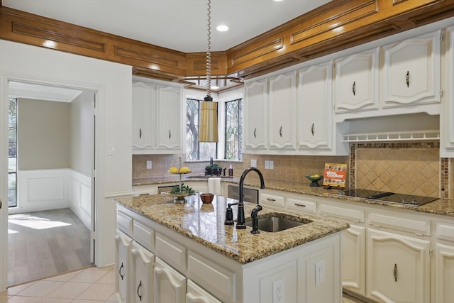 kitchen featuring light tile patterned floors, an island with sink, a sink, wainscoting, and black electric stovetop