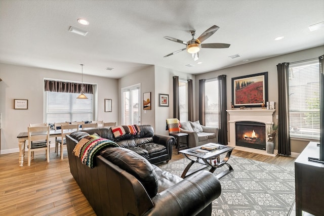 living room featuring a ceiling fan, baseboards, visible vents, light wood-style flooring, and a warm lit fireplace