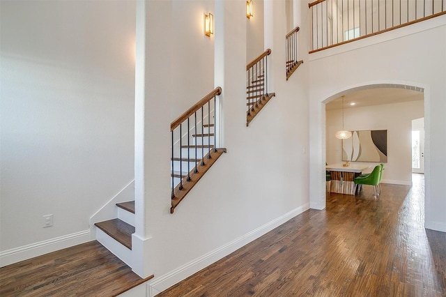 staircase with wood finished floors, arched walkways, a towering ceiling, and baseboards