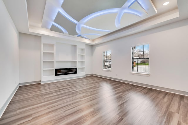 unfurnished living room with a raised ceiling, built in shelves, wood finished floors, and baseboards