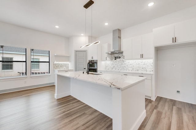 kitchen featuring light wood finished floors, backsplash, a center island with sink, white cabinetry, and wall chimney exhaust hood