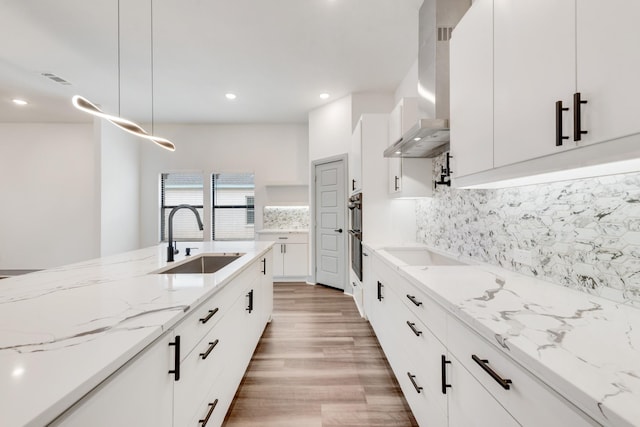 kitchen featuring wall chimney range hood, light wood finished floors, backsplash, and a sink