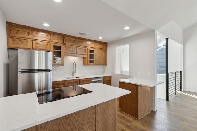 kitchen with wood finished floors, a sink, stainless steel appliances, glass insert cabinets, and brown cabinets