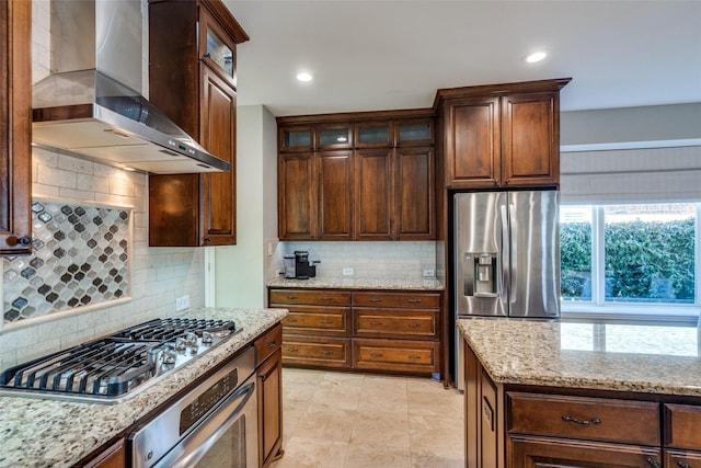 kitchen with light stone counters, stainless steel appliances, glass insert cabinets, wall chimney exhaust hood, and backsplash