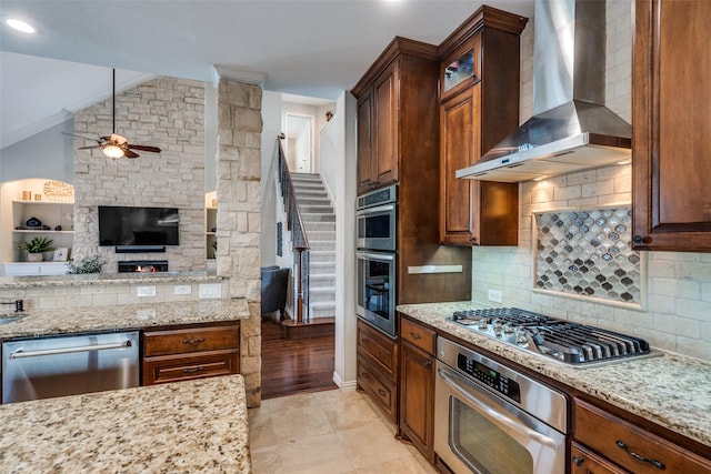 kitchen featuring wall chimney range hood, light stone countertops, open floor plan, decorative backsplash, and appliances with stainless steel finishes