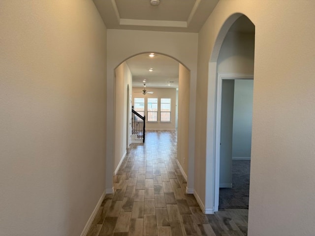 hallway featuring baseboards, stairway, a tray ceiling, wood finished floors, and arched walkways