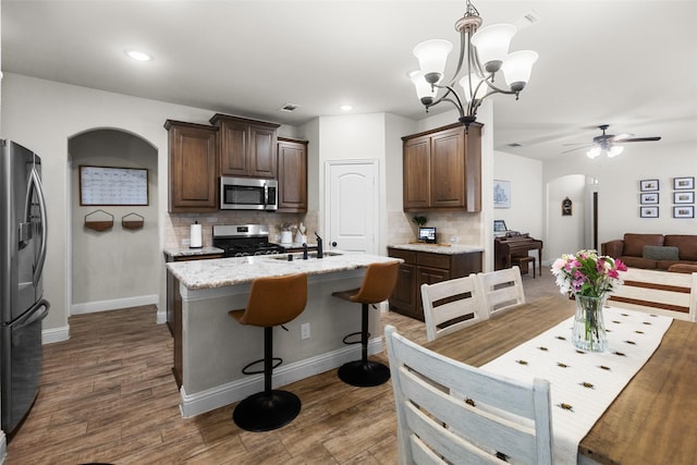 kitchen featuring a kitchen island with sink, arched walkways, dark brown cabinets, appliances with stainless steel finishes, and tasteful backsplash