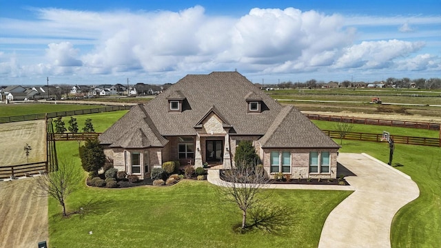 french provincial home with a front lawn, fence, a rural view, roof with shingles, and brick siding