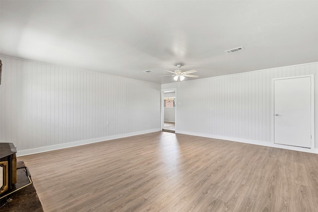 empty room with visible vents, light wood finished floors, baseboards, ceiling fan, and a wood stove