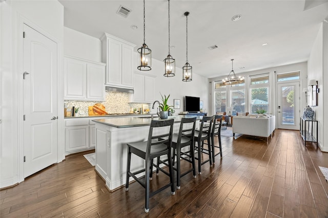 kitchen with an island with sink, dark wood-type flooring, white cabinets, under cabinet range hood, and backsplash