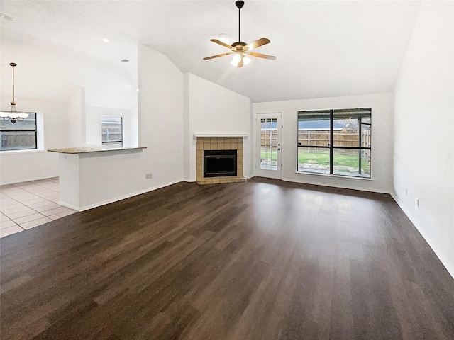 unfurnished living room featuring baseboards, high vaulted ceiling, dark wood-type flooring, a tiled fireplace, and ceiling fan with notable chandelier