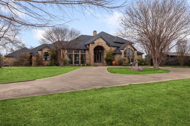 french country style house featuring a front lawn, roof with shingles, concrete driveway, brick siding, and a chimney