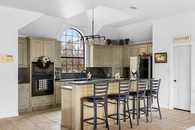 kitchen with a breakfast bar, an island with sink, vaulted ceiling, cream cabinetry, and black appliances