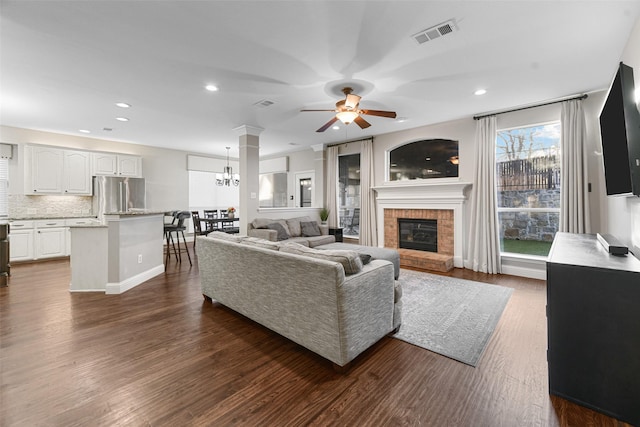 living area with dark wood-style floors, visible vents, recessed lighting, a brick fireplace, and ceiling fan with notable chandelier