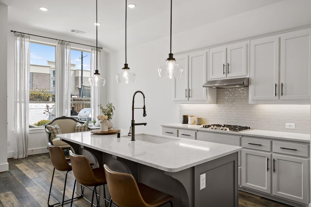 kitchen with a sink, stainless steel gas stovetop, under cabinet range hood, decorative backsplash, and dark wood-type flooring