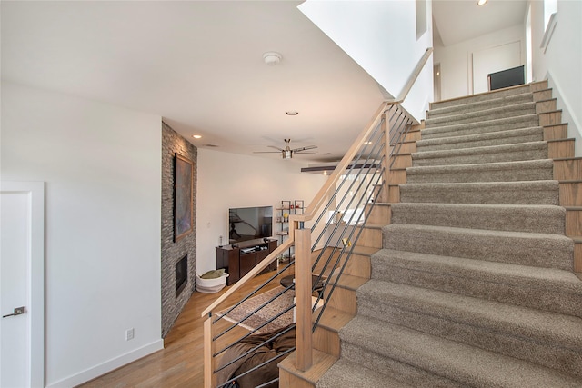 stairway featuring ceiling fan, wood finished floors, baseboards, and a large fireplace