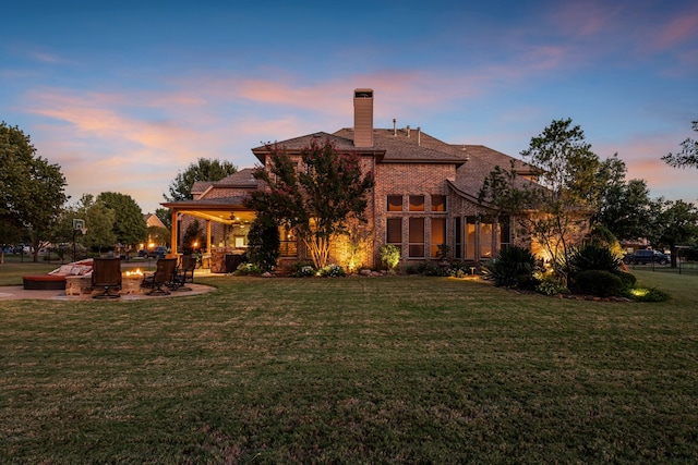 back of house at dusk with a yard, a chimney, brick siding, and a patio