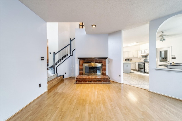 unfurnished living room featuring a ceiling fan, a textured ceiling, stairway, light wood-style floors, and a brick fireplace