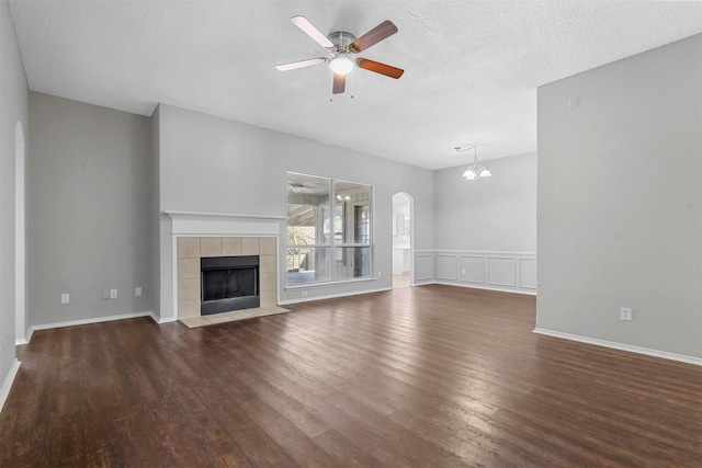 unfurnished living room featuring wood finished floors, arched walkways, a textured ceiling, a tiled fireplace, and ceiling fan with notable chandelier