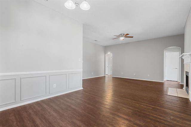 unfurnished living room with dark wood finished floors, arched walkways, a tile fireplace, a decorative wall, and ceiling fan with notable chandelier