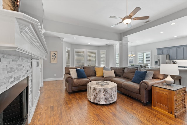 living area featuring wood finished floors, visible vents, ornate columns, a stone fireplace, and ceiling fan with notable chandelier