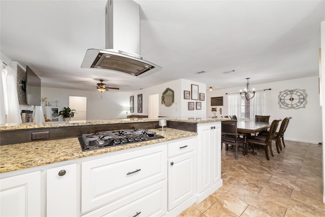 kitchen with light stone counters, island exhaust hood, ceiling fan with notable chandelier, white cabinets, and black gas stovetop