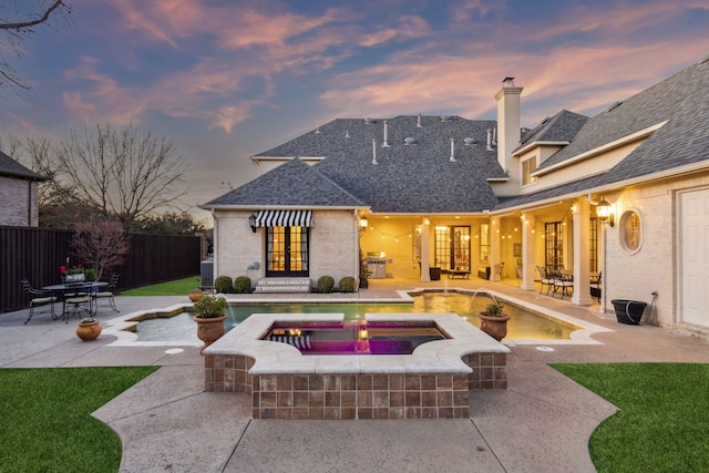 back of property at dusk with fence, roof with shingles, a chimney, a patio area, and brick siding