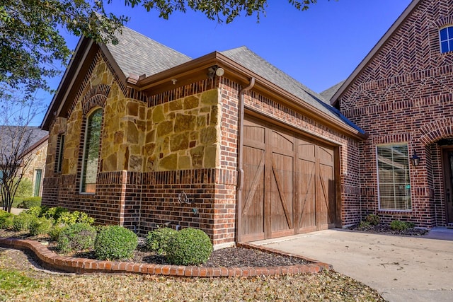 view of property exterior with driveway, a shingled roof, stone siding, a garage, and brick siding