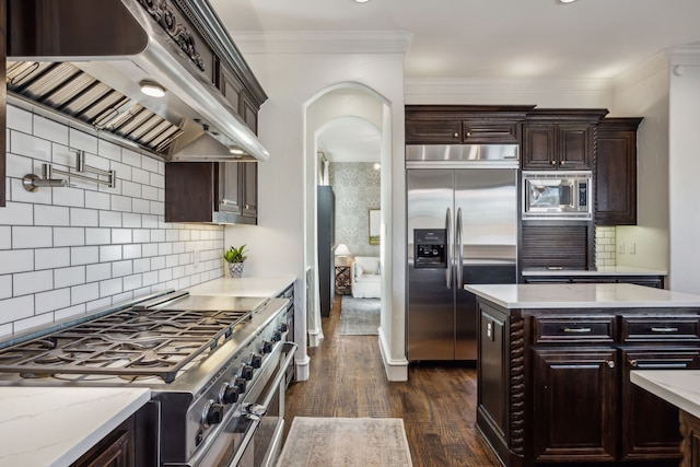 kitchen featuring ornamental molding, dark wood-type flooring, dark brown cabinetry, built in appliances, and under cabinet range hood