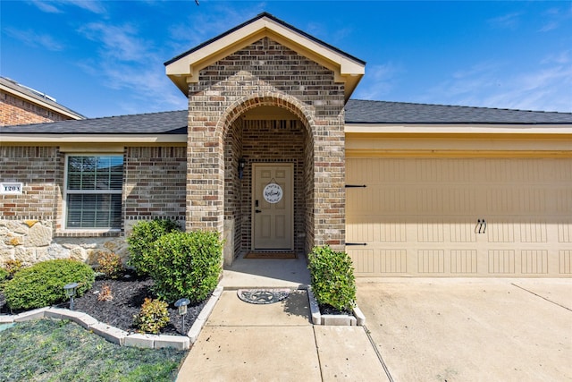 doorway to property featuring an attached garage, brick siding, driveway, and roof with shingles