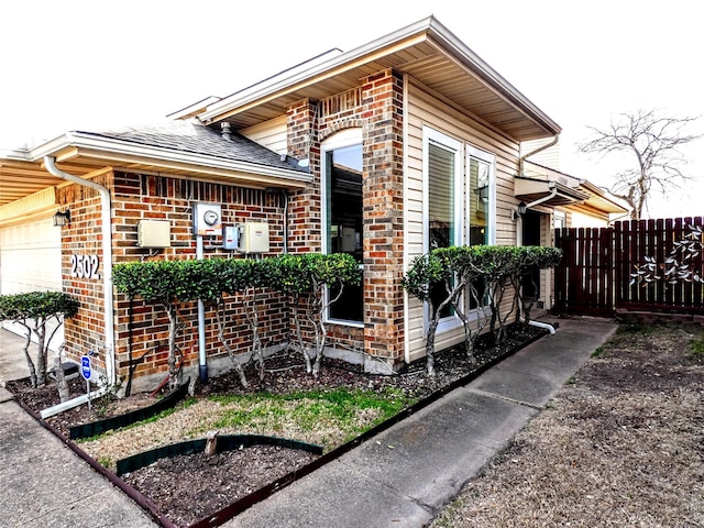 view of side of property with brick siding, roof with shingles, a garage, and fence