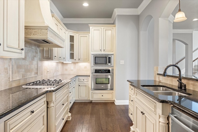 kitchen with dark stone countertops, custom exhaust hood, a sink, cream cabinetry, and appliances with stainless steel finishes