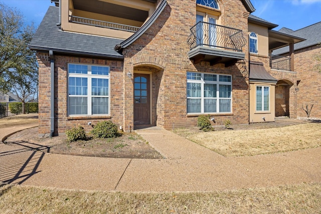 view of exterior entry with a balcony, brick siding, and roof with shingles