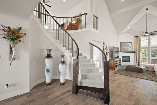 stairs with a large fireplace, baseboards, ceiling fan, wood finish floors, and high vaulted ceiling