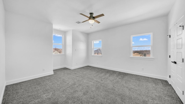 empty room with a wealth of natural light, a ceiling fan, visible vents, and dark carpet